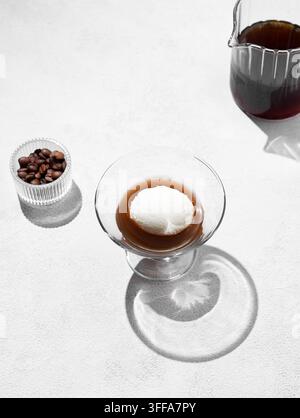 Affogato-Kaffee mit einer Kugel Vanilleeis und Espresso in einem Glas auf hellem Hintergrund mit Kaffeebohnen, Kanne und Schatten. Klassisches italienisches Des Stockfoto