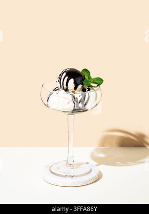 Löffel Vanilleeis in einem hohen Glas mit Schokoladenüberzug und einem Minzblatt auf einem hellen Hintergrund mit Schatten. Sommerliches erfrischendes Dessertkonzept Stockfoto