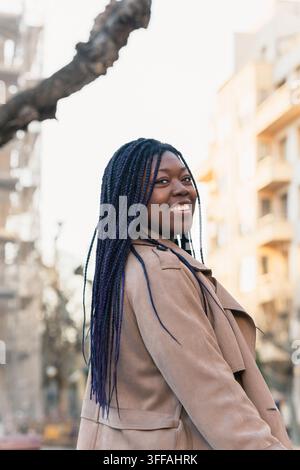 Porträt einer glücklichen schwarzen Frau mit langen Zöpfen, die auf einer urbanen Straße spaziert Stockfoto