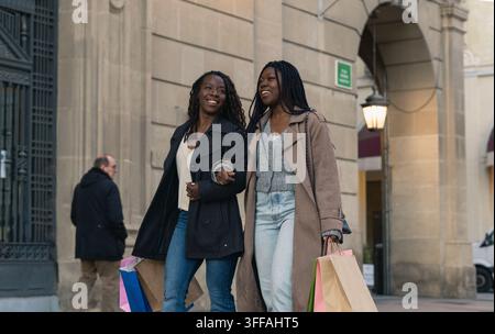 Zwei fröhliche afrikanische Freunde schlendern Arm in Arm nach dem Einkaufen an einem Frühlingsnachmittag. Stockfoto