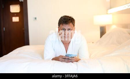 Mann in weißem Gewand liegt auf dem Bett und zählt Bargeld in Dollar – Konzept eines erfolgreichen Lebens. Stockfoto