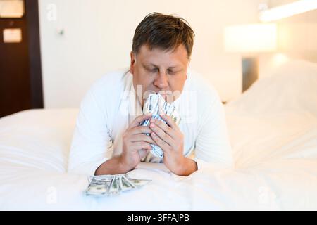 Mann in weißem Gewand liegt auf dem Bett und zählt Bargeld in Dollar – Konzept eines erfolgreichen Lebens. Er riecht den Duft des Geldes Stockfoto