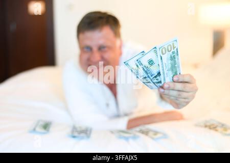 Mann in weißem Gewand liegt auf dem Bett und zählt Bargeld in Dollar – Konzept eines erfolgreichen Lebens. Selektive Fokussierung Stockfoto