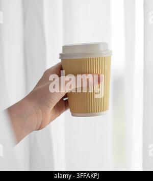 Hand, die einen Pappkaffeebecher mit weißem Deckel vor einem Fenster mit sanftem Tageslicht hält. Morgenroutine und To-go-Getränke-Konzept. Kopierraum. Stockfoto
