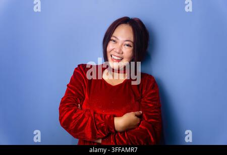 Fröhliche ostasiatische Frau mittleren Alters lacht mit verschränkten Armen, trägt ein rotes Samt-Top vor einem einfarbigen blauen Studiohintergrund. Stockfoto