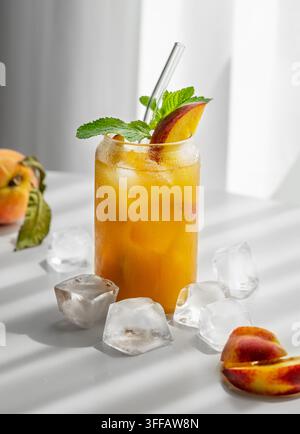 Erfrischendes Pfirsich-Cocktail, Limonade oder Eistee auf einem weißen Tisch in der Nähe des Fensters mit Schatten und Eiswürfeln. Konzept für einen kalten alkoholischen oder nicht-alkoholischen Drink Stockfoto