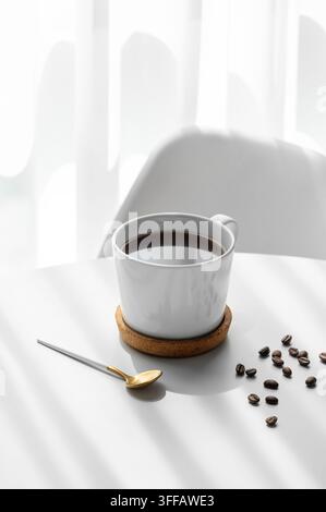 Frisch gebrühter Espresso in einer Tasse auf einem weißen Tisch nahe dem Fenster mit Löffel, Stuhl, Kaffeebohnen und Morgenlicht. Konzept einer Belebung Stockfoto