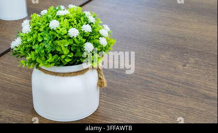 Tischdekoration im Café. Künstliche grüne Pflanze mit kleinen weißen Blüten in einem Keramiktopf steht auf einem Holztisch. Gemütliche Innenraum-Mittelstückkomposition. Stockfoto
