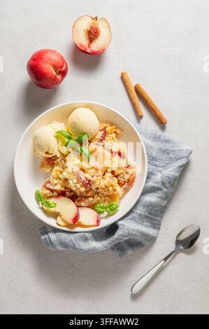Pfirsich-Crumble oder -Cobbler mit Vanilleeis-Scoops und Zimt auf einem Teller vor einem hellen Hintergrund mit frischen Früchten. Sommerliches hausgemachtes erfrischendes Dessert Stockfoto