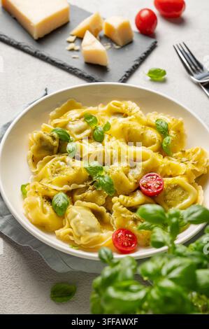 Tortellini-Pasta mit Parmesan, Pesto und Basilikum auf einem Teller vor einem hellen Hintergrund mit Tomate, Käse und Kräuter-Close-up. Traditionell italienisch hausgemacht Stockfoto