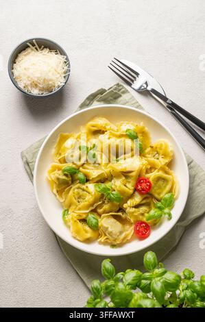 Tortellini-Pasta mit Parmesan, Pesto und Basilikum auf einem Teller vor einem hellen Hintergrund mit Käse, Kräutern und Besteck. Traditionelles italienisches Hausgemachtes Essen con Stockfoto