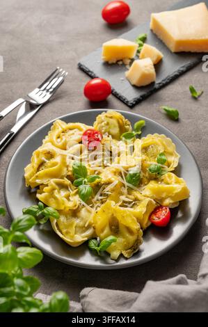 Tortellini-Pasta mit Parmesan, Pesto und Basilikum auf einem Teller vor dunklem Hintergrund mit Käse, Kräutern und Tomate. Traditionelles italienisches hausgemachtes Lebensmittelkonzept Stockfoto
