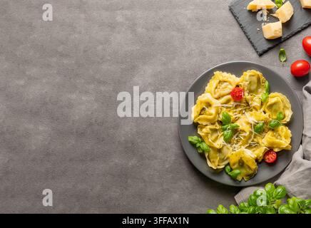 Tortellini-Pasta mit Parmesan, Pesto und Basilikum auf einem Teller vor dunklem Hintergrund mit Käse, Kräutern und Tomate. Traditionelles italienisches hausgemachtes Lebensmittelkonzept Stockfoto