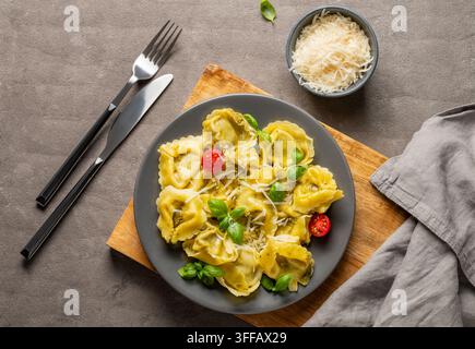 Tortellini-Pasta mit Parmesan, Pesto und Basilikum auf einem Teller auf einem Holzbrett auf einem dunklen Hintergrund mit Käse und Besteck. Traditionell italienisch hausgemacht Stockfoto