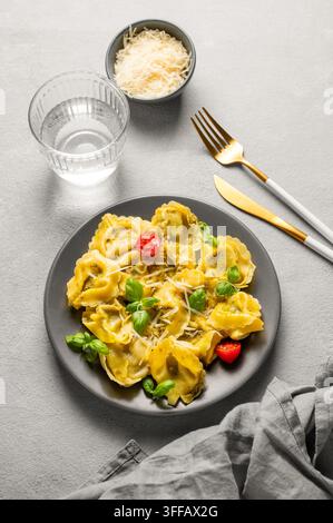 Tortellini-Pasta mit Parmesan, Pesto und Basilikum auf einem Teller vor einem hellen Hintergrund mit einem Glas Wasser, Käse und Besteck. Traditionell italienisch hausgemacht Stockfoto