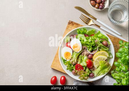 Thunfischsalat mit Oliven, Salat, Tomaten, Eiern und Zitrone auf einem Teller auf einem Holzbrett auf einem hellen Hintergrund mit einem Glas Wasser und Besteck. Die Konc Stockfoto