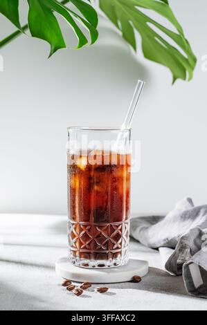 Kaltes Brew-Kaffee-Tonikum in einem Glas mit Eis auf einem hellen Hintergrund mit Kaffeebohnen, Monstera-Blatt und Morgenschatten. Sommerliche, erfrischende Trend-Begleitung Stockfoto