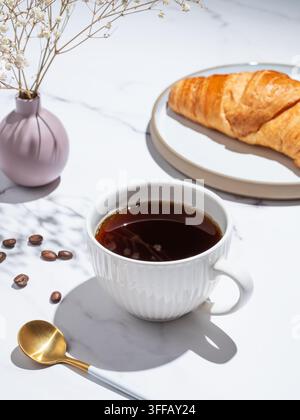 Frisch gebrühter schwarzer Kaffee in einer Tasse und Croissant auf einem Teller auf einem weißen Marmorhintergrund mit Löffel, Blume und natürlichem Morgenlicht. Konzept von Stockfoto