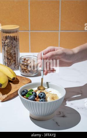 Hand hält einen Löffel in griechischem Joghurt mit Granola, Blaubeere und Banane in einer Schüssel auf einem Marmortisch mit Obst und Morgenlicht. Gesund und nahrhaft Stockfoto