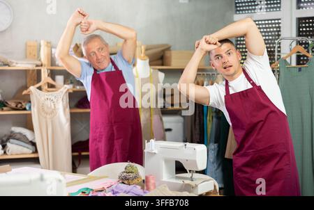 Männliche Schneider in roten Schürzen stehen in der Werkstatt und wärmen sich auf. Nähmeister machen während einer Pause Muskeldehnübungen Stockfoto