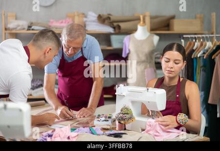 Männliche Schneider stehen neben einem Tisch und zeichnen eine Skizze von Kleidung neben einer weiblichen Schneiderin, die an einer Maschine arbeitet. Team von Modedesignern und Schneidern Stockfoto
