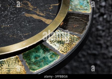 Makrofotografie eines dekorativen Mosaiktischrandes mit glasierten Keramikfliesen, einem polierten Messingrand und einer strukturierten schwarzen Tischplatte. Natürliches Crackle p Stockfoto