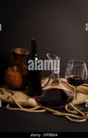 Dramatische Rotwein-Stillleben mit einem vollen Dekanter, Glas, Flasche und Tonkrug, gruppiert mit Sackleinen und Seil vor einem tiefschwarzen Hintergrund. Stockfoto