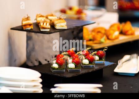 Elegante Vorspeisen auf gestuften schwarzen Schiebetabletts arrangiert. Kirschtomaten, Mozzarella und Basilikum garnieren kleine Glasbecher. Goldene Backwaren sitzen auf Holz Stockfoto