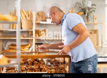 Seniorer männlicher Kunde bleibt am Fenster stehen und schaut sich Produkte in den Regalen des Ladens an. Kunde wählt Gebäck in der Bäckerei, überlegt zu kaufen. Brötchen und croi Stockfoto