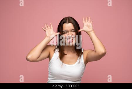 Junge Frau, die die Zunge herausstreckt und die Hände am Gesicht hält, die Verspieltheit und Humor auf einem rosa Hintergrund ausdrückt Stockfoto