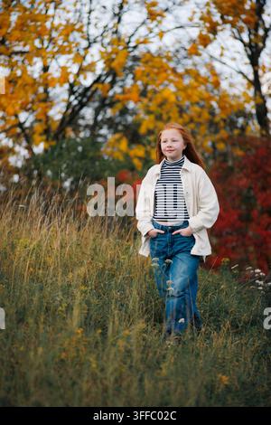 Vollständige Aufnahme eines rothaarigen Mädchens, das durch ein Wiesenfeld mit hohem, trockenem Gras geht, mit verschwommenem, leuchtend gelbem und rotem Herbstlaub der Bäume Stockfoto