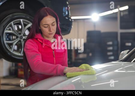 Eine Frau reinigt ein Auto mit einem Tuch in einer Autowerkstatt tagsüber. Sie konzentriert sich darauf, die Oberfläche des Fahrzeugs zu reinigen. Stockfoto