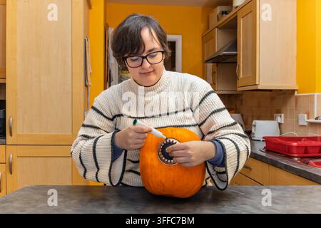 Nahaufnahme einer Frau in gestreiftem Pullover, die einen Metallabflusssieb als Schablone gegen einen orangefarbenen Kürbis hält und mit einem grünen Marker einen Kreis nachzeichnet, um sich vorzubereiten Stockfoto