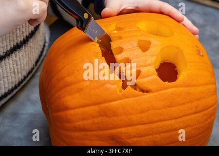 Nahaufnahme von Händen, die mit einem Küchenmesser einen Mund in eine große orangefarbene Halloween-Kürbisse schneiden, Gesichtsmerkmale bereits teilweise ausgehöhlt, hohles Innere Stockfoto