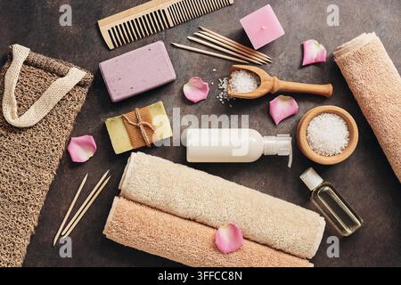 Flat Lay eines Sets verschiedener Badaccessoires. Frottierhandtuch, Seife, Kamm, Öl, Shampoo, Luffa-Waschhandschuh, Meersalz auf dunklem rustikalem Hintergrund. Blick von oben Stockfoto