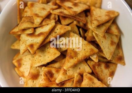 Eine Aufsichtsfotografie, die einen vollen, dichten Haufen goldbrauner dreieckiger gebackener Pita-Chips zeigt, die mit Kräutern und Gewürzen gewürzt sind, in einer weißen Schüssel. Stockfoto