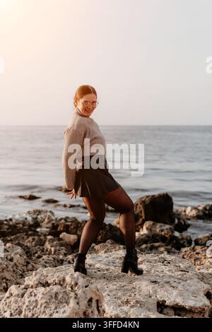 Frau am Meeresufer, glückliche Frau in Pullover und schwarzem Rock steht an einer felsigen Küste und blickt beim Sonnenuntergang zurück Stockfoto