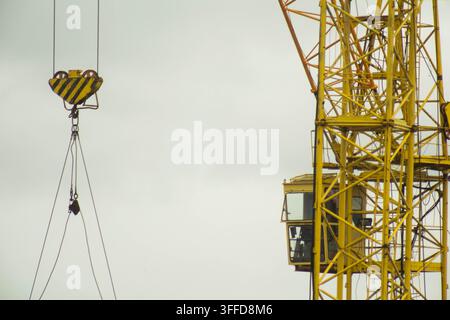 Gelbe Turmdrehkran-Struktur mit Kabine für den Bediener vor bewölktem Himmel. Haken, gestreift in Schwarz und Gelb, hängt von Stahlseilen. Maschinerie für er Stockfoto