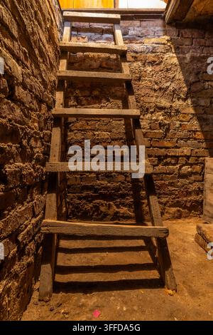 Alte Holzleiter lehnt an einer rauen Ziegelwand in einem schwach beleuchteten Keller. Staubige Umgebung mit Schmutz und Trümmern auf dem Boden verstärkt die rustikale Atmosphäre. Stockfoto