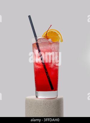 Eleganter roter Cocktail mit Eiswürfeln, serviert in einem Glas auf einem Sockel, dekoriert mit einer frischen Orangenscheibe und einer Kirsche vor einem hellen Hintergrund. Erfrischend Stockfoto
