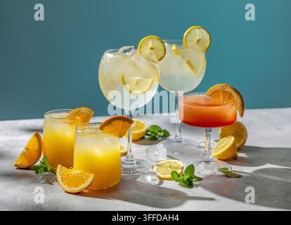 Sortiment von farbenfrohen erfrischenden Getränken, serviert mit Eis und garniert mit frischer Orange, Zitrone, Limette und Minze auf einem sonnenbeschienenen grauen Tisch. Helle Sommer Stockfoto