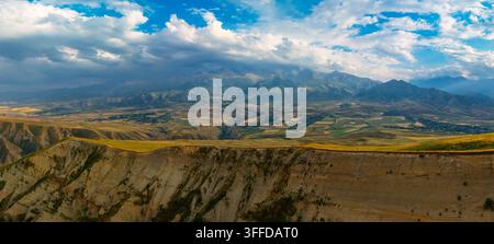 Panoramische Luftaufnahme zeigt eine raue Berglandschaft mit einer steilen Felswand und einem breiten Tal darunter. Sanfte Hügel und ein kleines Dorf sind unter einem dynamischen Himmel mit dramatischen Wolken und Sonnenlicht sichtbar. Stockfoto
