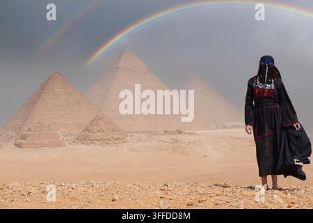 Traditionell verschleierte ägyptische Frau in kunstvollem Volkskostüm steht auf dem sandigen Giza-Plateau und blickt auf die antiken Pyramiden unter einem atemberaubenden Doppel Stockfoto