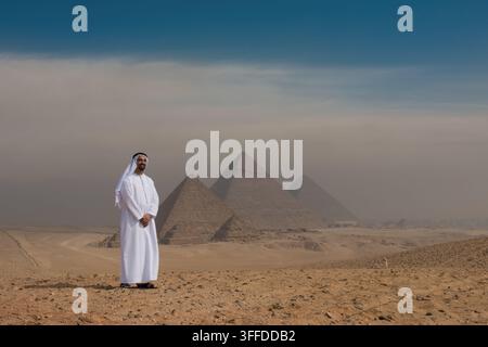 Stolzer Emirati in weißer Kandura und Keffiyeh steht lächelnd vor allen drei Pyramiden von Gizeh in Ägypten mit atmosphärischem Wüstennebel und offenem Blau Stockfoto