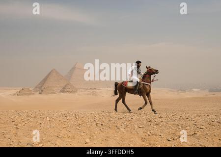 Selbstbewusster arabischer Mann auf einem Araberpferd, der über das Wüstengebiet von Gizeh galoppiert, während alle drei großen Pyramiden im Hintergrund unter einem diesigen bewölkten Himmel stehen Stockfoto