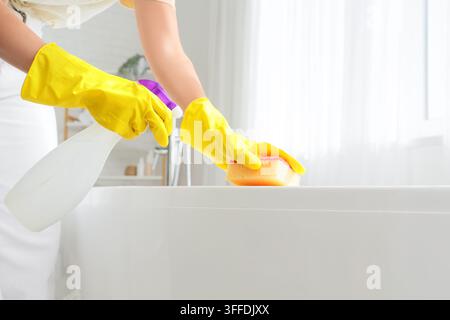 Hausreinigung und Hygiene-Wartung, Person trägt gelbe Gummihandschuhe und verwendet eine Sprühflasche und einen Schwamm, um eine weiße Badewanne im Badezimmer zu desinfizieren Stockfoto