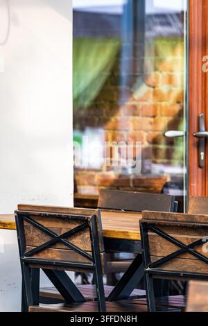Gemütliche Sitzgelegenheit im Außenbereich eines Cafés mit Holzstühlen mit X-Rücken aus Metallrahmen und einem Massivholztisch. Warmes Sonnenlicht wirft weiche Schatten gegen ein Glas Stockfoto
