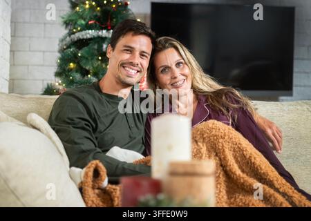 Lächelndes romantisches Paar, das eng beieinander unter einer warmen Decke auf dem Sofa sitzt und einen gemütlichen Weihnachtsmoment zu Hause mit einem geschmückten Baum genießt Stockfoto