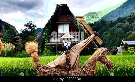 verspielte traditionelle Vogelscheuche steht in einem lebendigen grünen Reisfeld vor einem steilgiebeligen Gasshō-zukuri-Bauernhaus in Shirakawa-go. Stockfoto