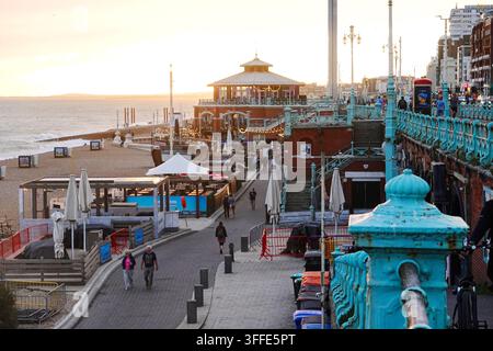 Menschen schlendern entlang der unteren Strandpromenade an Strandcafés vorbei bei Sonnenuntergang in Brighton, England. Stockfoto